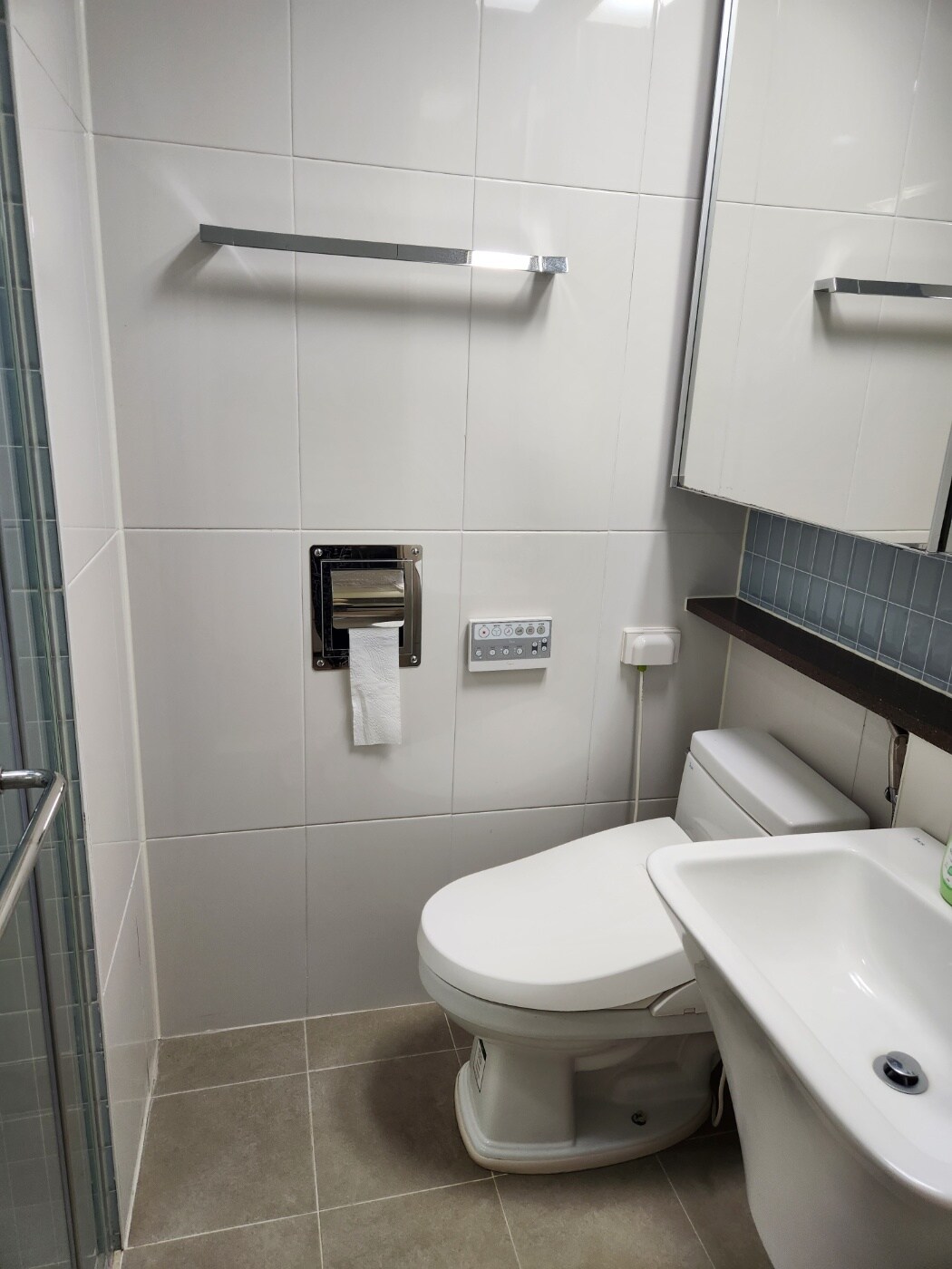The bathroom features modern fixtures including a toilet and a sink with a sleek counter. A mirror is mounted above the sink, and a towel holder is located on the wall. The space is finished with clean, white tiles and soft lighting.