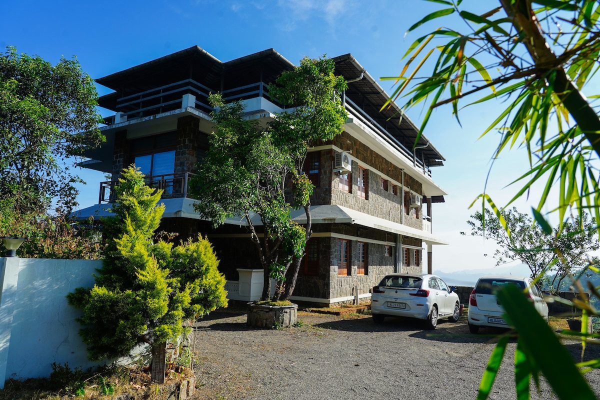 The villa is set amidst nature, showcasing a three-story structure with a mix of stone and modern architecture. Surrounding greenery complements the exterior, while parking spaces are visible in front. The layout emphasizes a connection with the outdoors and offers panoramic views of the landscape.