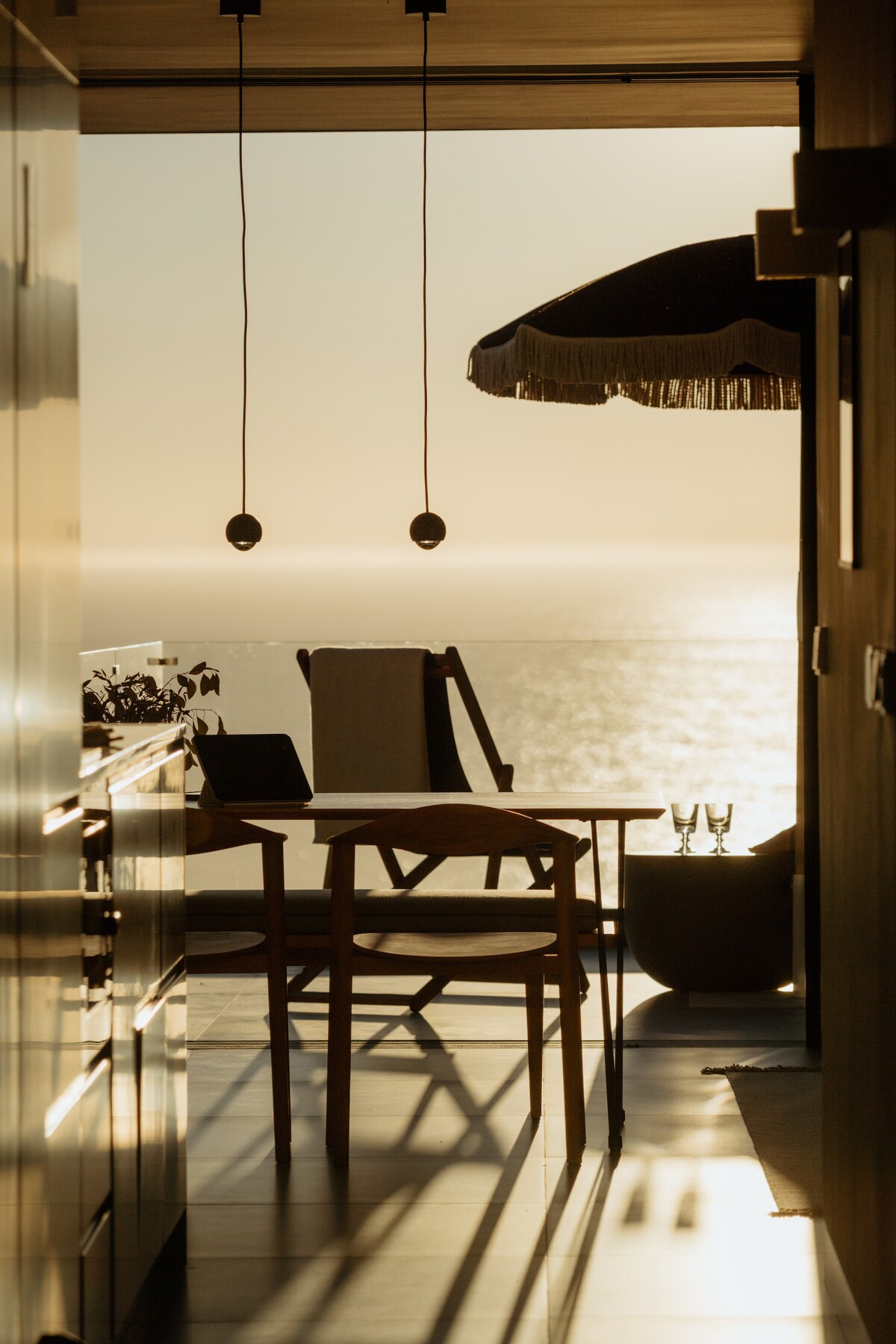 A dining area is highlighted by warm light, showcasing a wooden table with a chair. A terrace with a sunshade and seating overlooks the shimmering ocean beyond. The glow of sunset creates soft shadows, enhancing the intimate atmosphere of the space.
