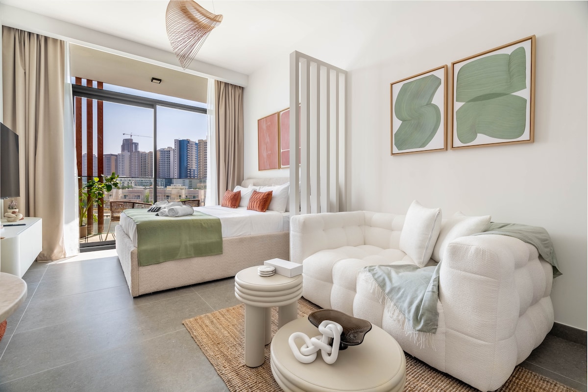 An open-plan living area features a comfortable sofa and a spacious bed dressed in soft linens. Natural light floods the room through large windows, enhancing the relaxing atmosphere. Decorative artwork adorns the walls, contributing to the modern aesthetic.