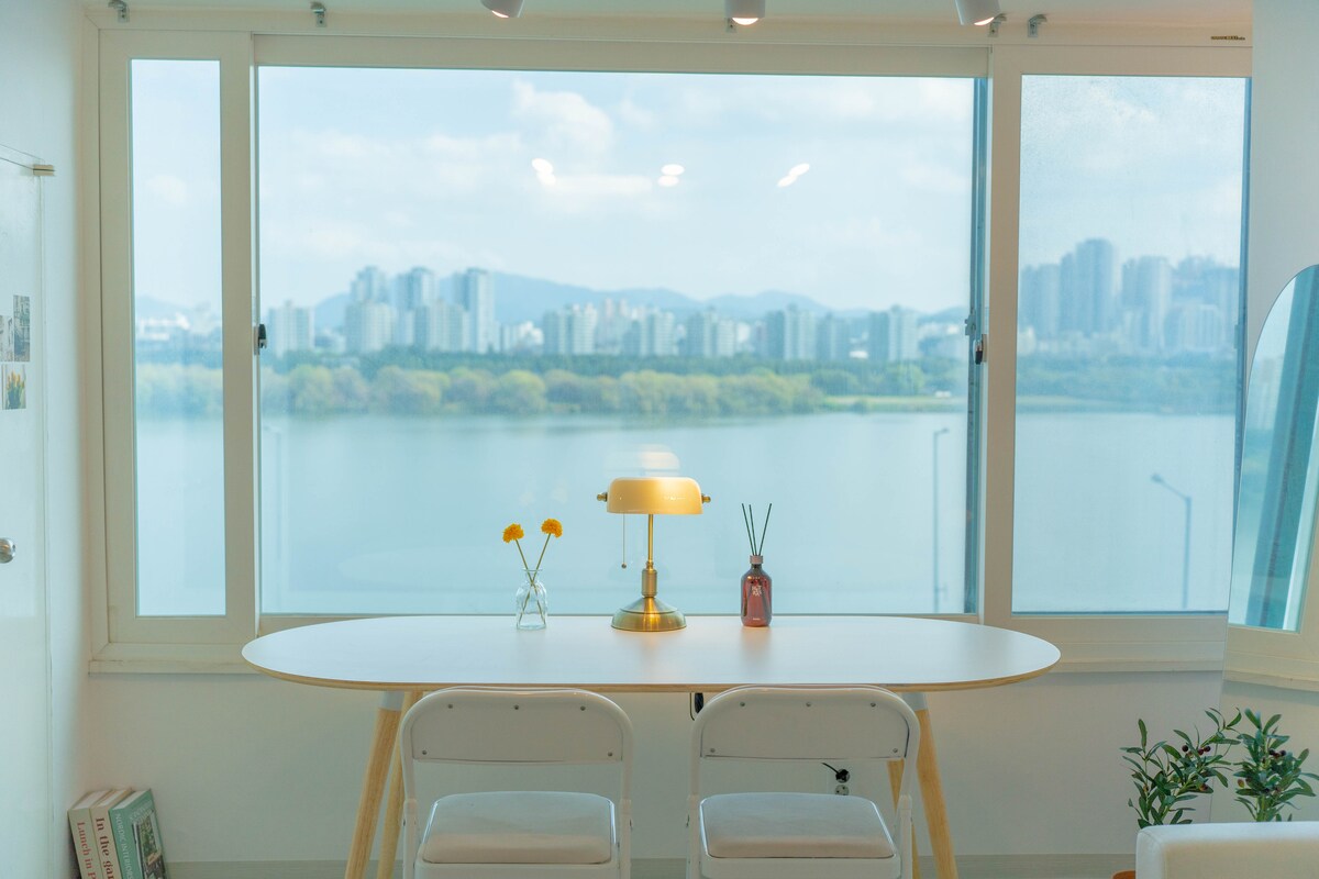 A dining table is positioned in front of a large window, overlooking a scenic view of the river and city skyline. Decorative elements include a lamp and two vases with flowers on the table. Natural light illuminates the space, enhancing the serene ambiance.