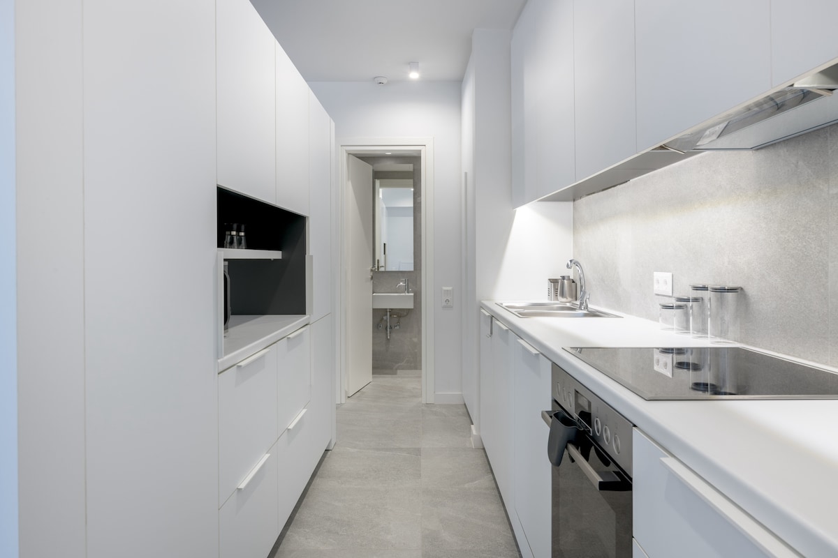 A modern kitchen with sleek white cabinetry is displayed, featuring a smooth countertop and built-in appliances. The well-organized space leads to a doorway that opens into a bathroom area, enhancing its functionality.
