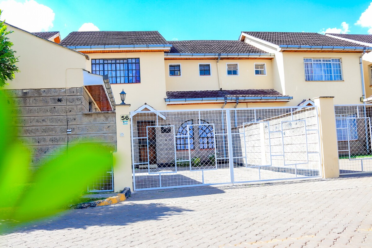The exterior of the home is framed by a secure gated entrance, showcasing a spacious front yard. A well-maintained façade features a combination of light-colored walls and a tiled roof. The driveway is bordered by greenery, contributing to the welcoming atmosphere.