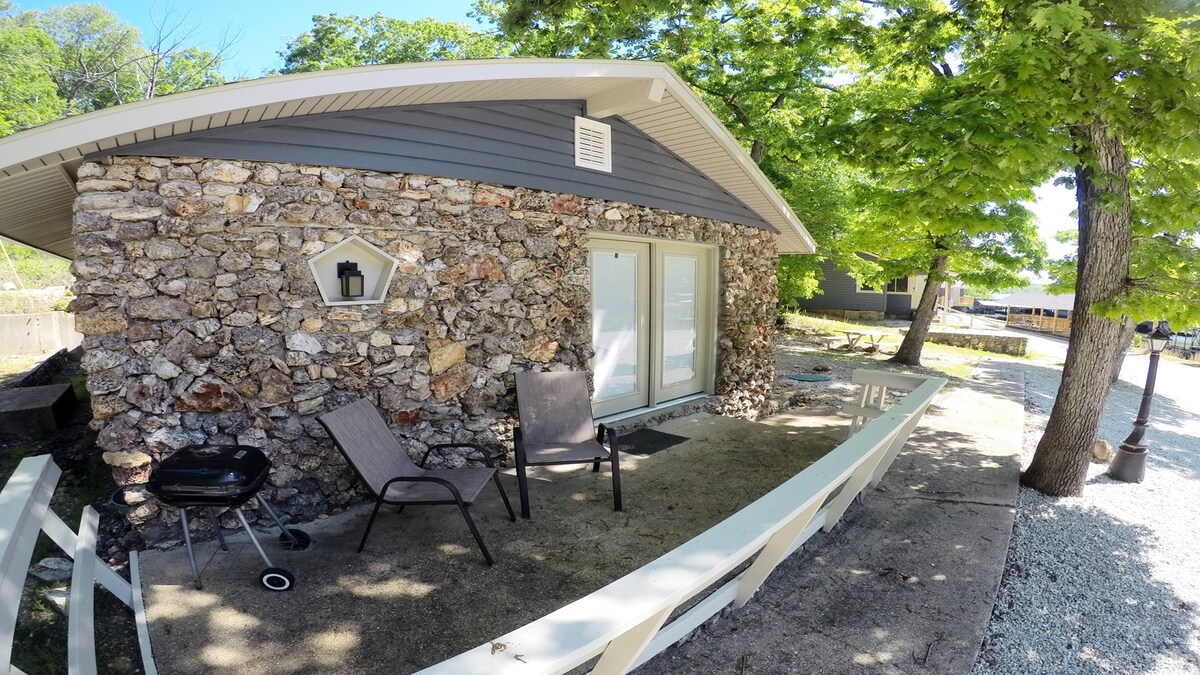 A rustic cabin exterior displays a stone façade surrounded by lush greenery. A small outdoor space features two lounge chairs and a BBQ grill, situated near a concrete area. Large sliding glass doors provide access to the interior, while a clear view of the natural surroundings is presented.