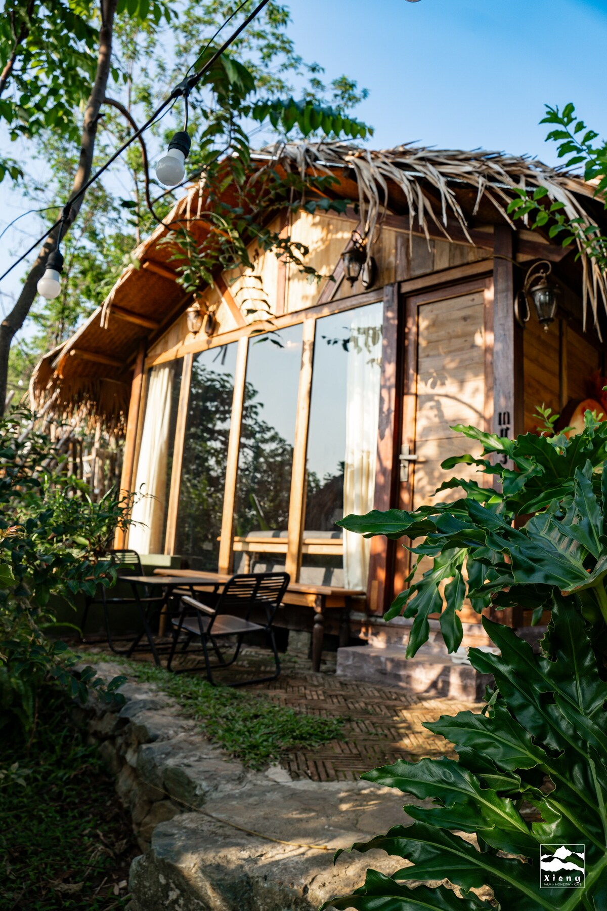 The wooden bungalow is framed by lush greenery, featuring large glass windows that invite natural light. A small outdoor seating area with two chairs and a table is positioned on a stone pathway, enhancing the connection to the surrounding garden.