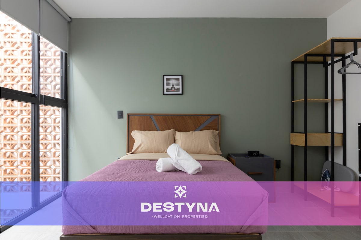 A cozy bedroom features a double bed with neatly arranged linens and a pair of rolled towels. Large windows allow natural light to fill the space, while a minimalist shelving unit and clothing rack provide storage. A muted green wall complements the room's decor.