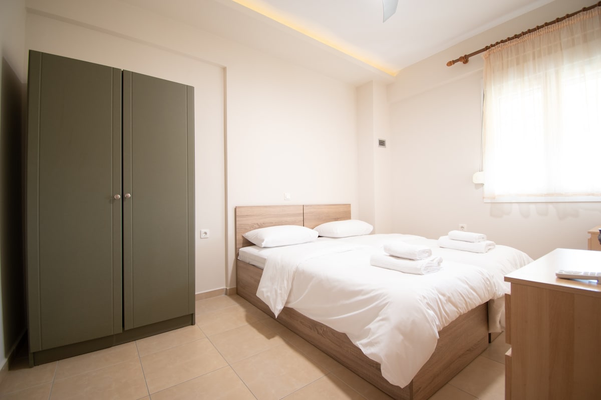 A bright and airy bedroom is presented, featuring a wooden bed with white linens and neatly arranged towels. A spacious wardrobe stands against the wall. Natural light filters in through a window covered by sheer curtains, creating a serene environment.