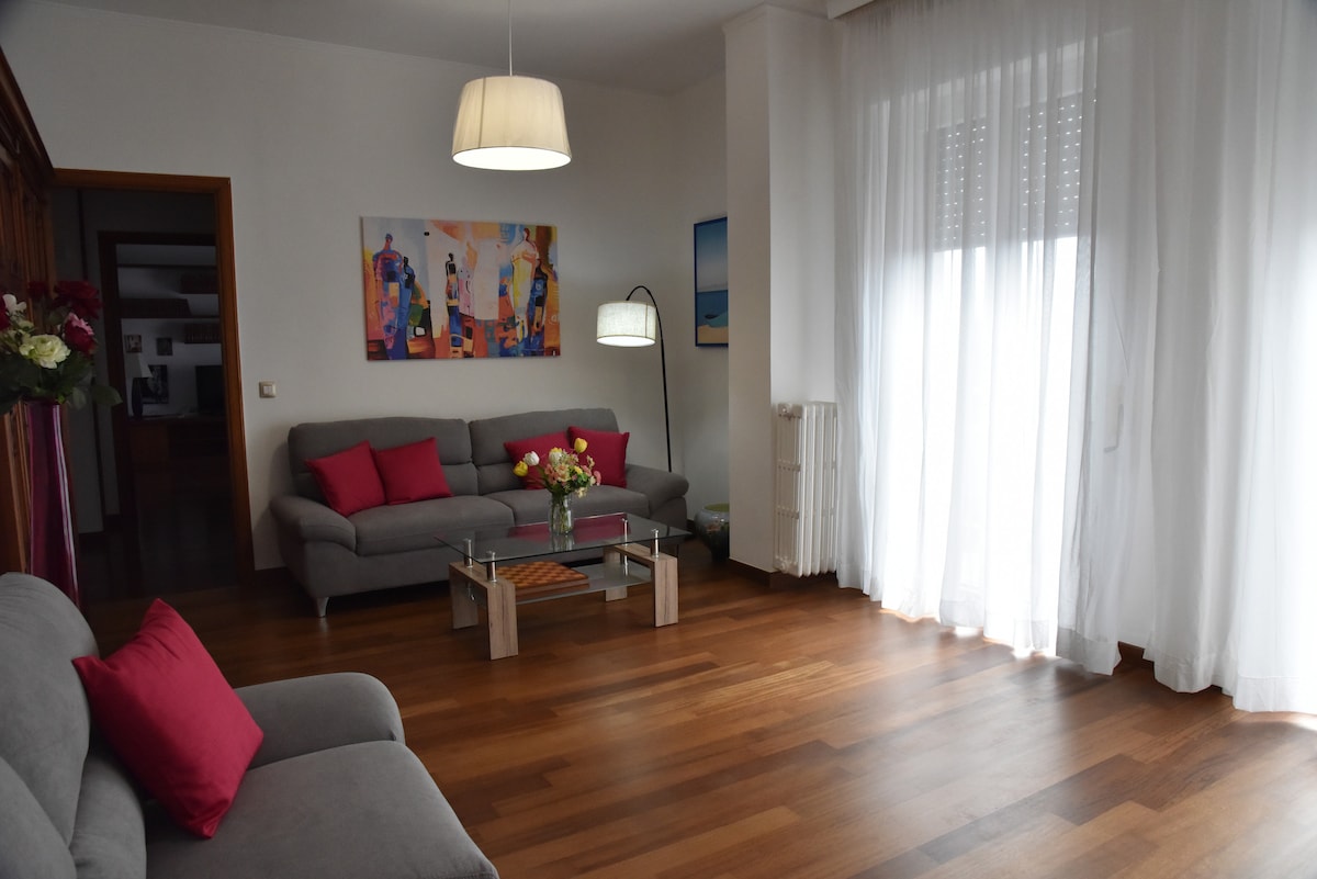 A bright living area features two light gray sofas adorned with red accent pillows, creating a comfortable seating arrangement. A glass coffee table sits in the center, surrounded by natural wood flooring. Large windows are dressed with sheer white curtains, allowing ample daylight to illuminate the space.