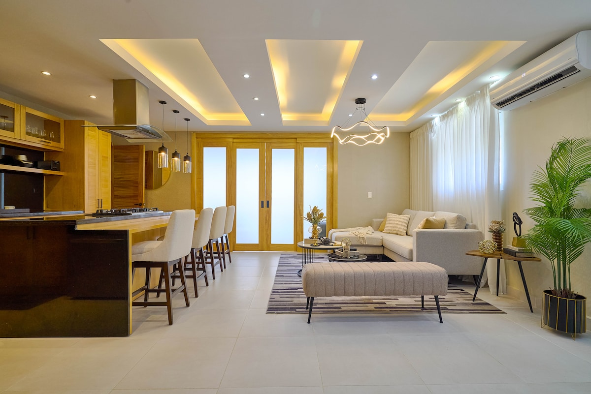 The spacious living area features a modern kitchen with a breakfast bar and seating for six. A light-colored sofa and accent chairs complement the neutral decor. A stylish coffee table and indoor plants add character, while large glass doors enhance natural light.