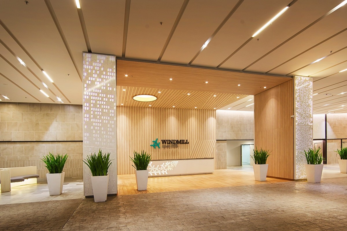 A modern, well-lit lobby area is depicted, featuring a combination of smooth wooden panels and elegant stone walls. Large potted plants are positioned on either side of the entrance, enhancing the welcoming appearance of the space. The building’s name 'Windmill' is prominently displayed.