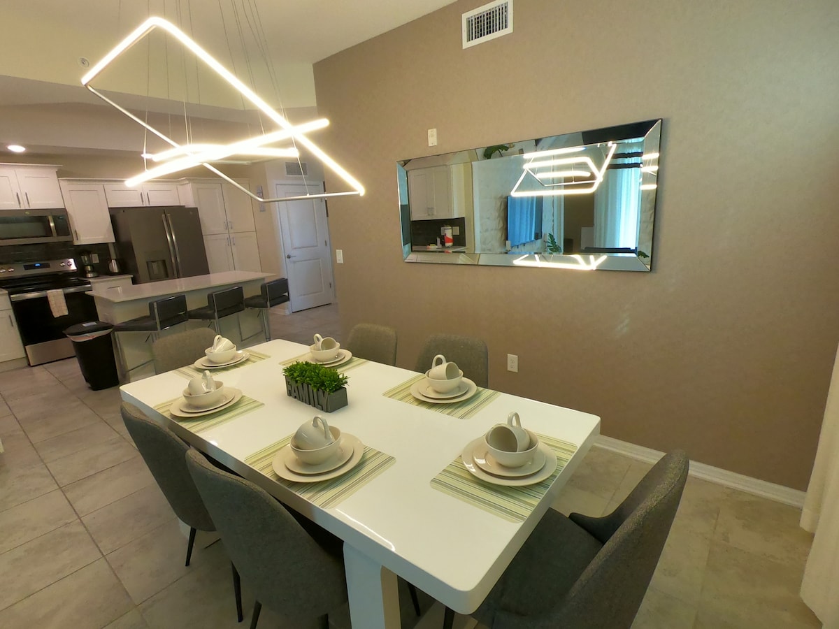 A modern dining area showcases a white rectangular table set for six, featuring elegant dishware and placemats. A contemporary light fixture hangs overhead, complemented by a large mirror reflecting the room's design. Neutral walls and tile flooring provide a cohesive backdrop.