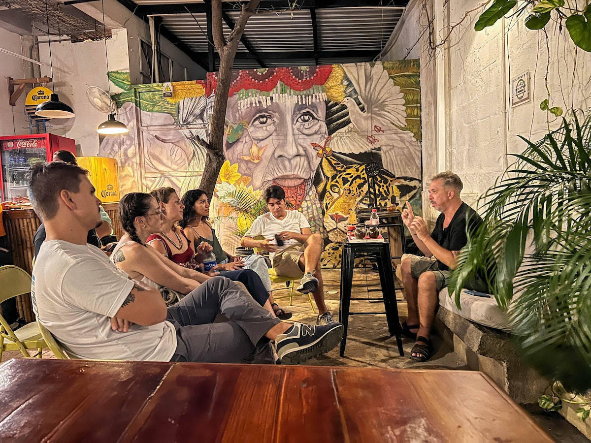 A casual gathering is depicted in a vibrant communal space, featuring a striking mural of a face on the wall. Guests are seated on various chairs, engaging in conversation, with plants in the foreground adding a touch of greenery to the atmosphere.