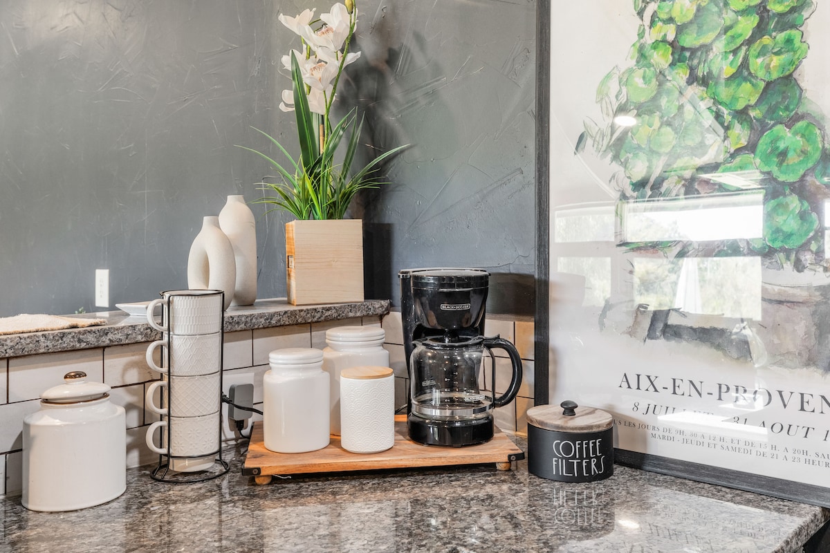 A well-organized kitchen counter features a coffee maker, various storage jars, and decorative elements. A plant adds a touch of greenery, while a framed artwork provides an aesthetic backdrop. The counter is made of textured granite, contributing to a functional and inviting environment.