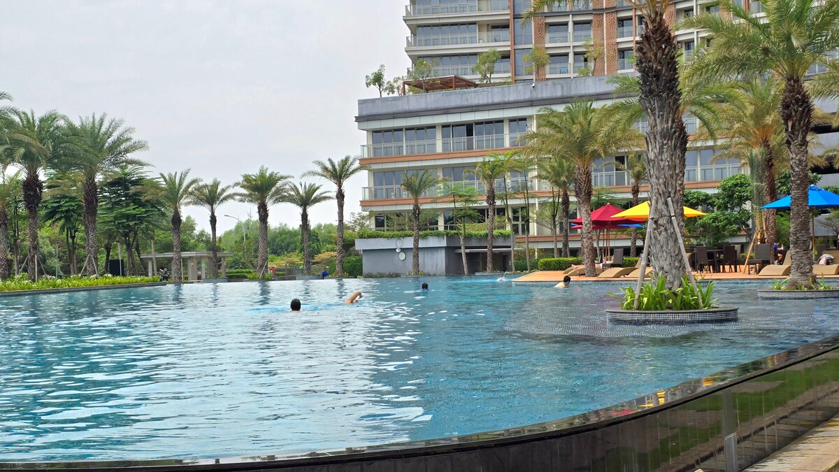 An expansive outdoor swimming pool is surrounded by palm trees and lounge chairs in vibrant colors. Guests can be seen enjoying the water, while the modern building stands in the background, contributing to the relaxed atmosphere.