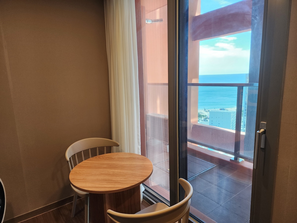 A small balcony area features a round wooden table with two light-colored chairs. Floor-to-ceiling glass doors offer a view of the ocean and distant buildings, with sheer curtains allowing natural light to filter in.