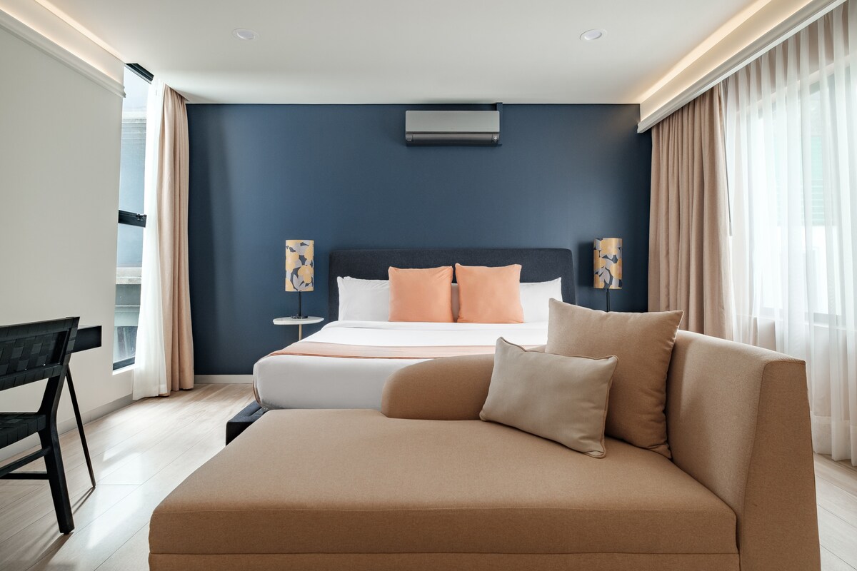 A modern suite features a spacious bed with soft linens, accompanied by two decorative lamps on either side. A cozy sofa with pillows provides comfortable seating, while large windows are dressed in light, airy curtains allowing natural light to fill the room.
