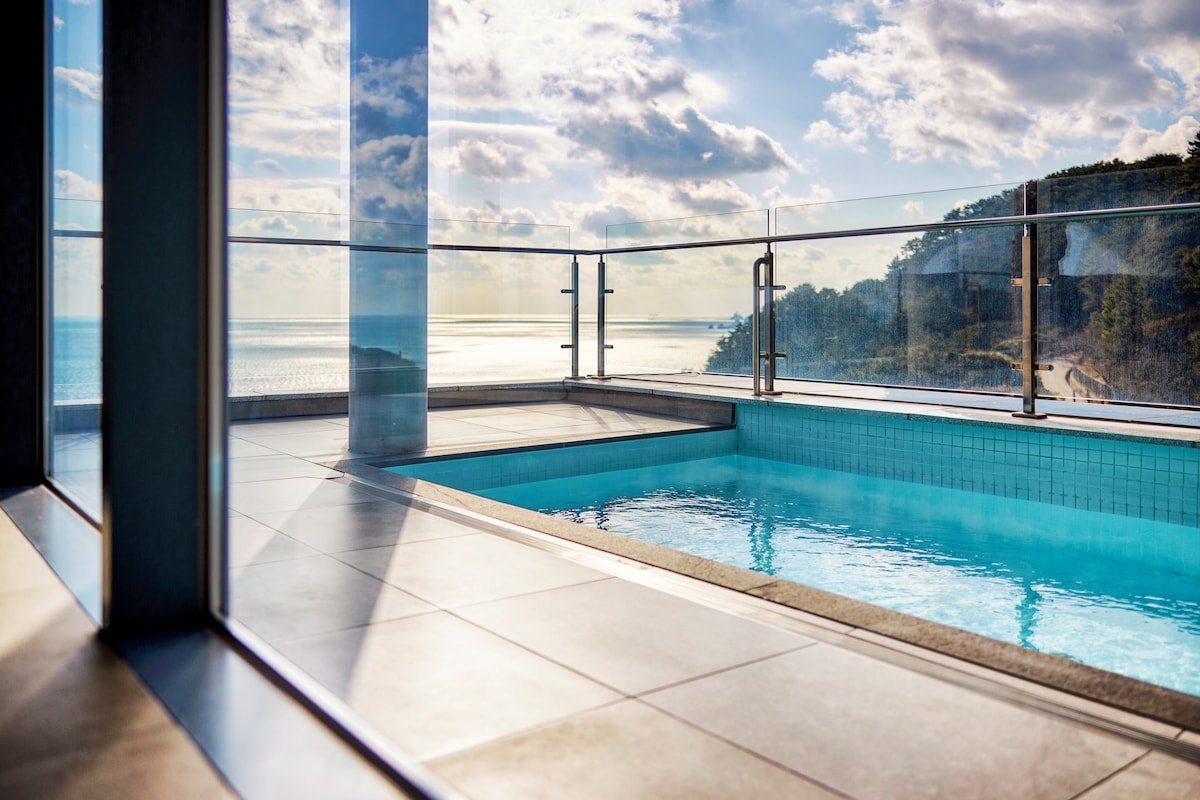A stunning outdoor pool with clear water is framed by glass panels, offering unobstructed views of the sea and sky. Sunlight reflects off the water surface, while the surrounding deck features modern tiles, emphasizing the pool's serene atmosphere.