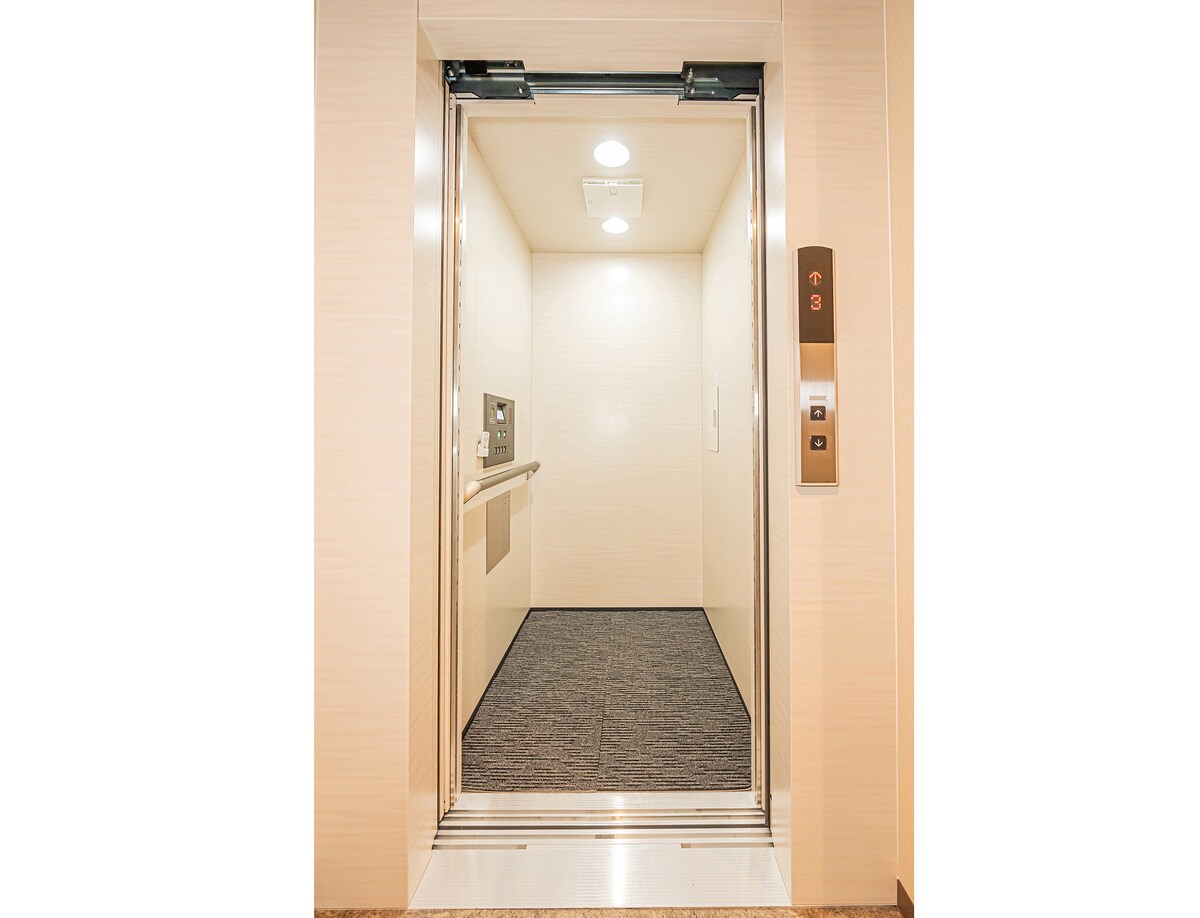An elevator entrance is shown, featuring smooth walls and a clean, spacious interior. The door is opened, revealing a neatly maintained floor with muted carpeting. An electronic control panel is visible next to the door, allowing convenient access to different floors.