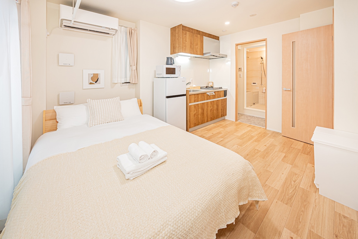 A bright and cozy room features a spacious double bed with soft bedding. A small kitchenette is equipped with essential appliances, including a microwave and refrigerator. A private bathroom is visible in the background, while wooden flooring adds a warm touch to the space.