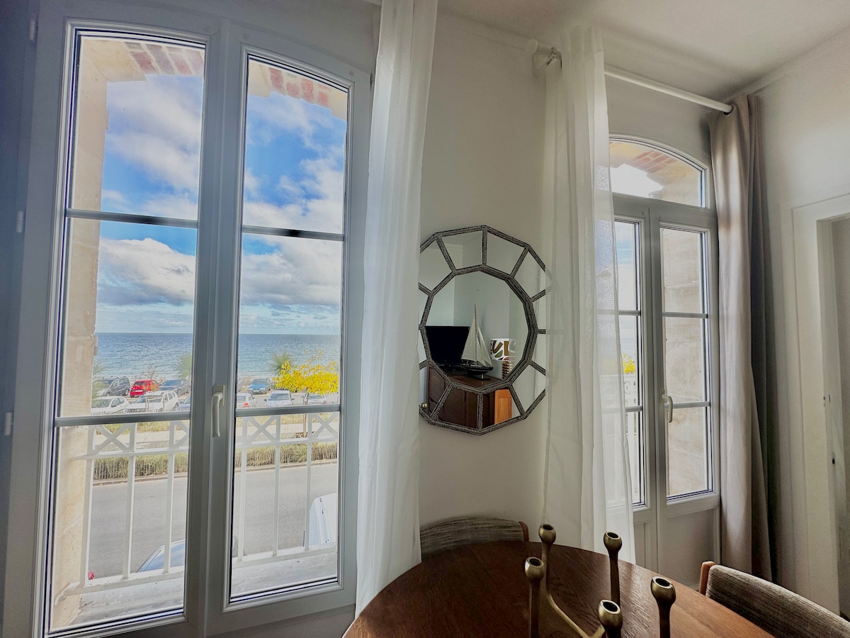 Large windows allow natural light to fill the room, offering a direct view of the sea. A round mirror hangs on the wall, reflecting the serene surroundings. A wooden dining table is positioned in the foreground, creating a welcoming atmosphere for meals.