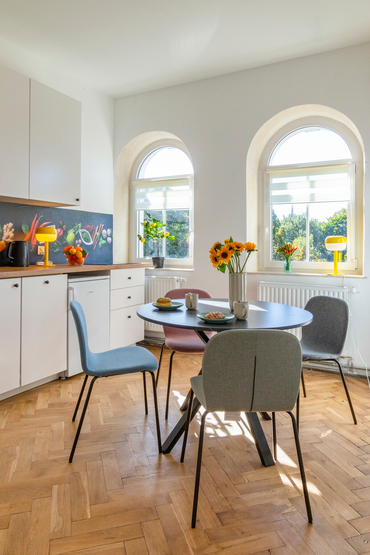 A bright kitchen area features a round dining table set for four, complemented by two light blue chairs and two gray chairs. Fresh flowers are placed in a vase on the table. Large arched windows allow natural light to fill the space, enhancing the cheerful atmosphere.