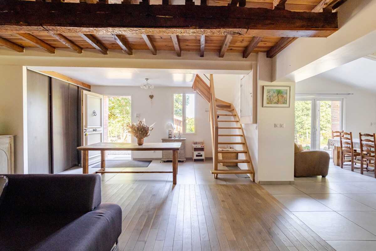 A spacious living area is featured, showcasing a wooden ceiling and a staircase leading to the upper level. A central table is surrounded by natural light, highlighting a cozy lounge area and an open kitchen corner. Large windows provide views of the greenery outside.