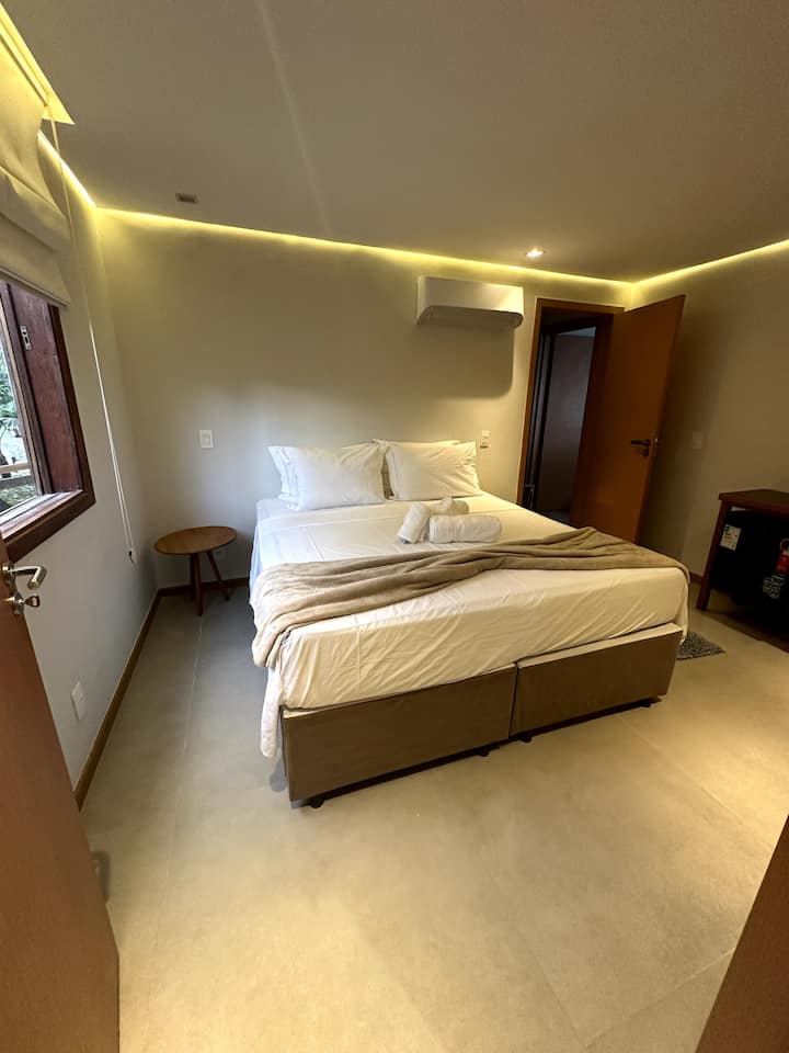 "Quarto luminoso e aconchegante com uma cama queen size, iluminação suave e decoração minimalista. Perfeito para uma estadia relaxante em Maraú, Bahia."