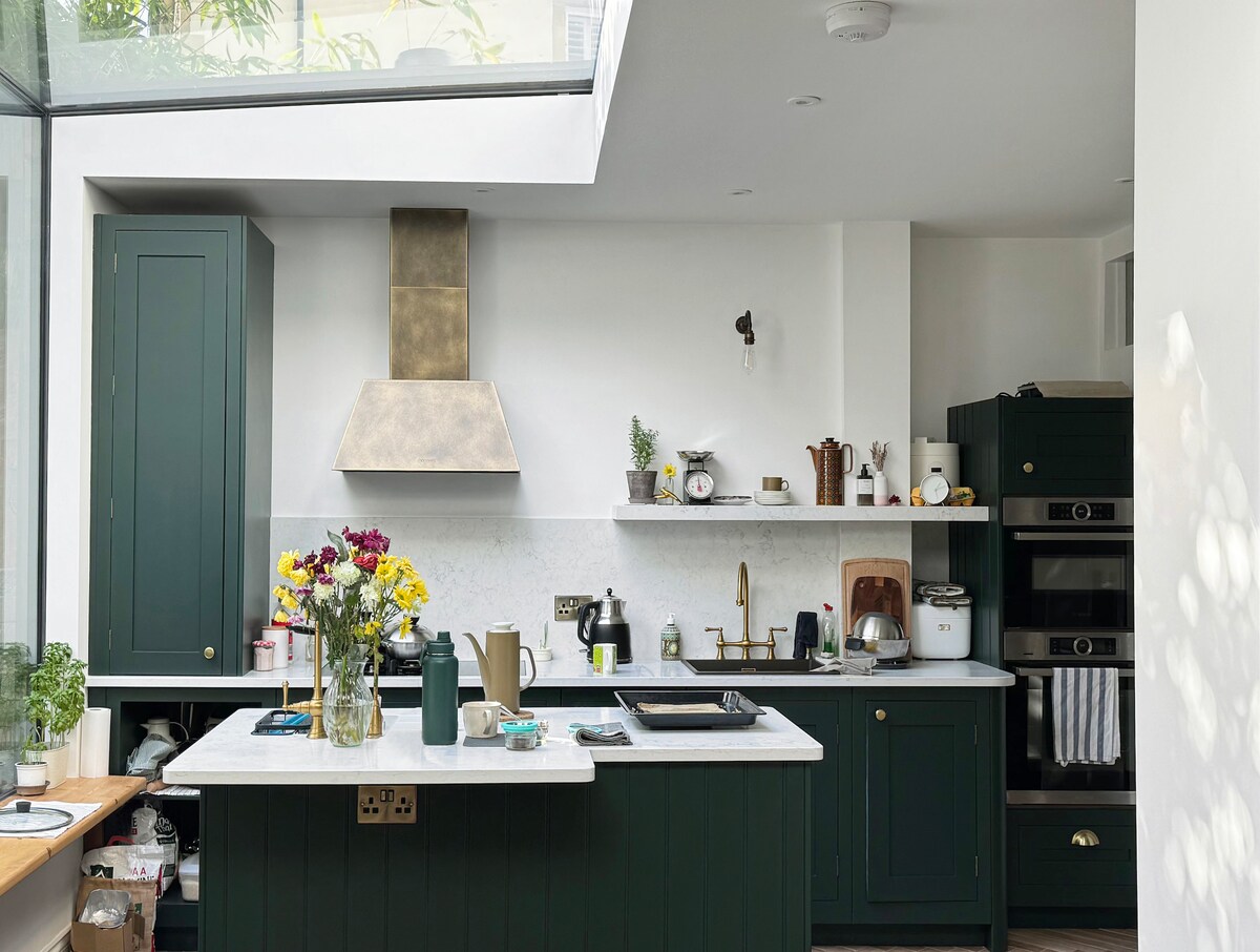 A modern kitchen with deep green cabinetry and a marble countertop is showcased. A stainless steel hood hangs above the stove, with an open shelf displaying various kitchen utensils. A bouquet of flowers adds a touch of color, while large windows allow natural light to fill the space.