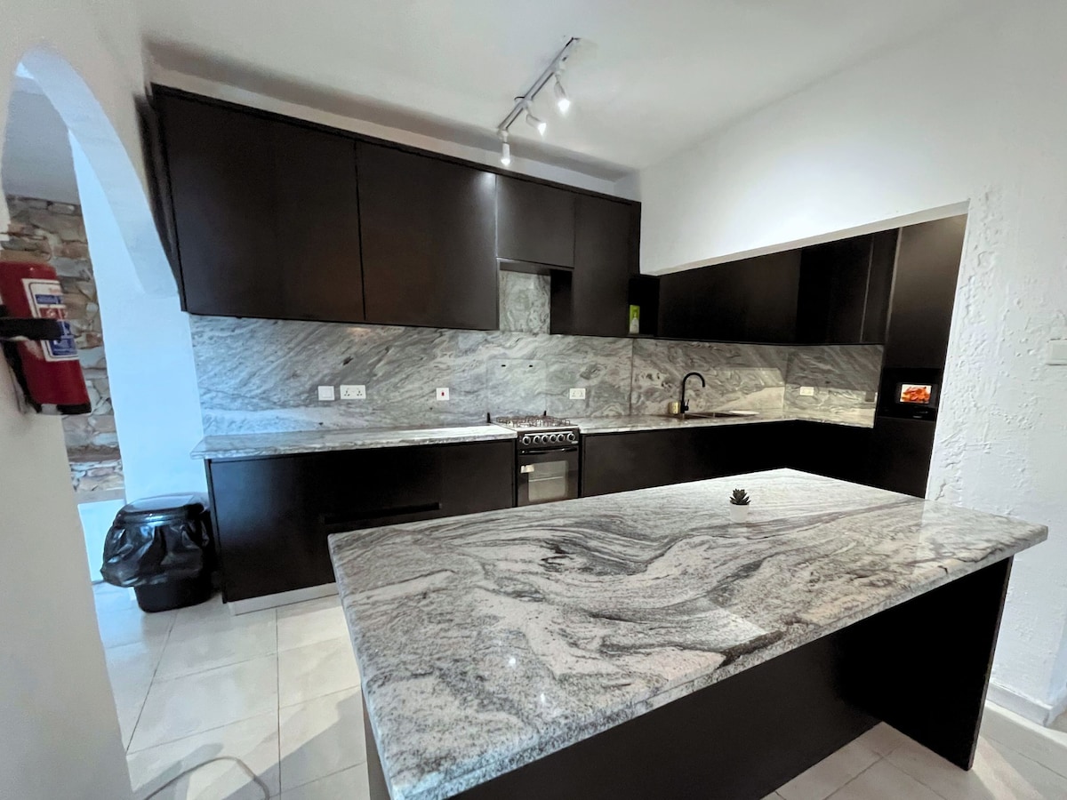 A modern kitchen features dark cabinetry complemented by a striking granite countertop. The kitchen is equipped with a stove and a sink, and under-cabinet lighting highlights the stone's texture. A spacious island provides additional workspace and a central gathering area.