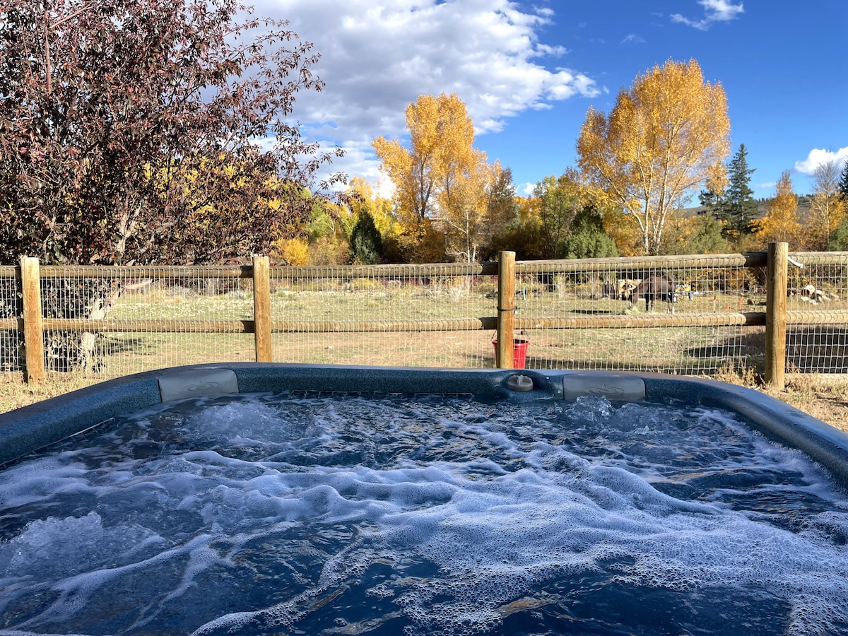 A hot tub is situated in a fenced area, with bubbling water visible. Surrounding trees display vibrant autumn foliage, while a clear blue sky enhances the serene setting. The landscape features a grassy field in the background, creating a peaceful outdoor retreat.