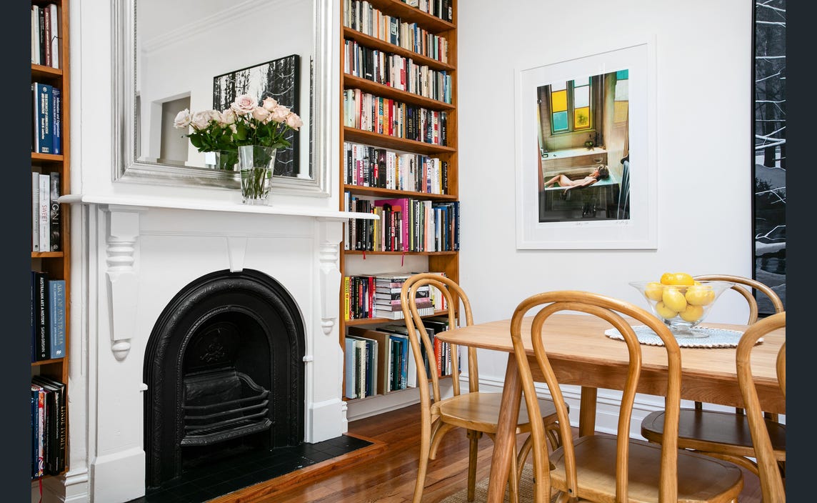 A dining area features a wooden table surrounded by chairs, positioned near a classic fireplace. Built-in shelves line one wall, filled with books. A bouquet of roses sits in front of a mirror, while a framed artwork adds a touch of color to the space.