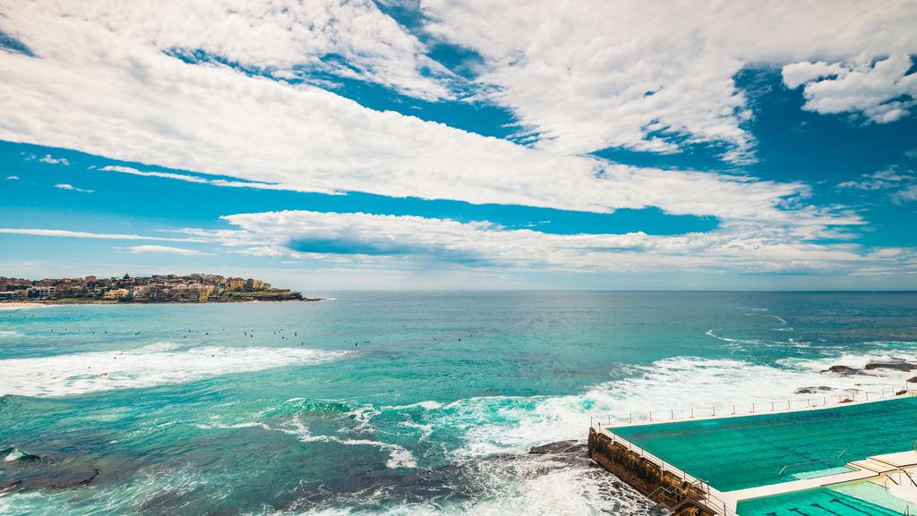 An expansive view of Bondi Beach showcases rolling waves meeting the shore. The vibrant turquoise water contrasts with the soft clouds above, creating a serene beachfront scene. The coastline is visible in the distance, providing a sense of the area’s natural beauty.