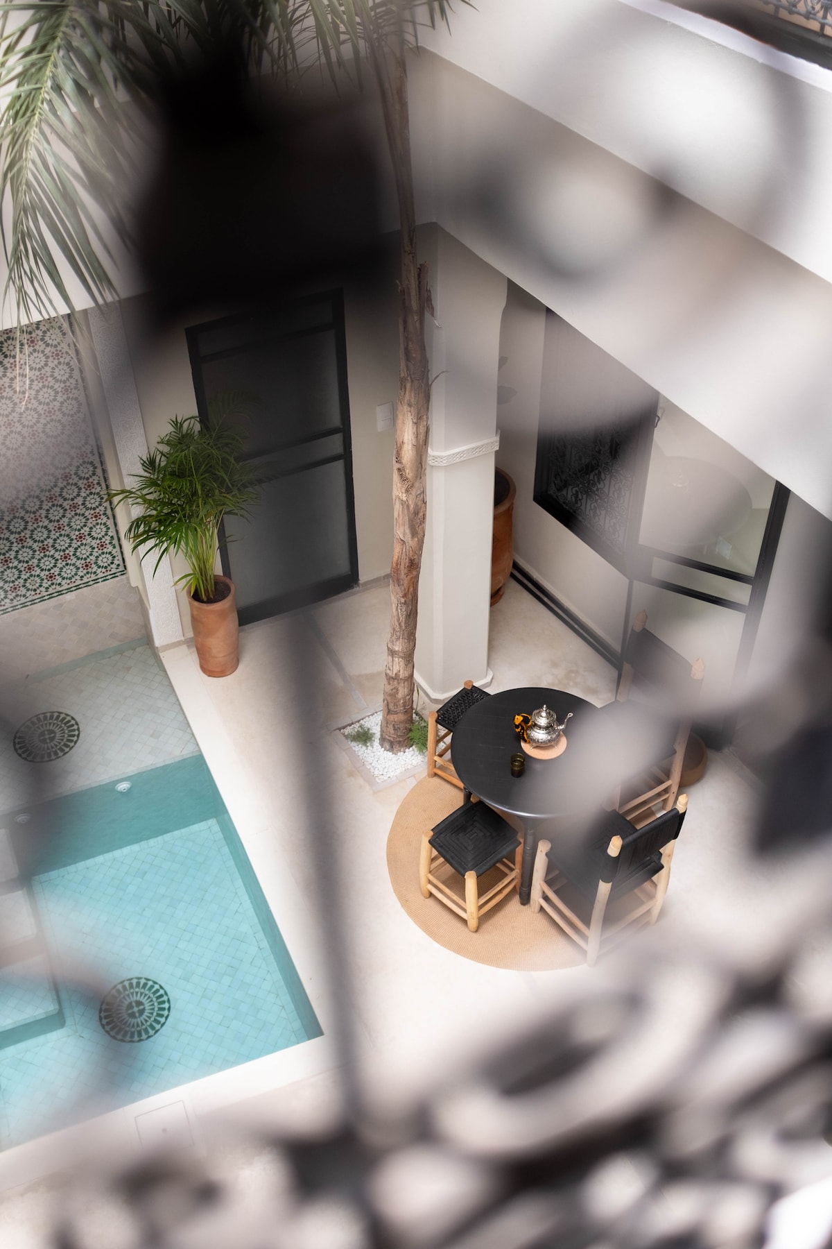A view of the riadâs serene courtyard, featuring a refreshing turquoise pool and a circular dining area equipped with black chairs surrounding a wooden table. Tall green plants add a touch of nature, and intricate tile patterns accentuate the space.
