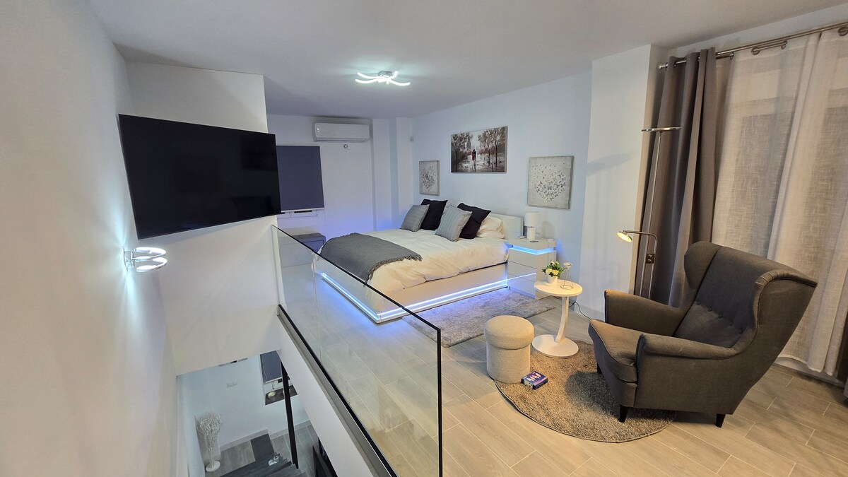 A modern loft area is highlighted with a spacious bed adorned with neutral bedding and decorative pillows. A large window allows natural light to brighten the space, complemented by elegant glass railings. A comfortable chair is positioned nearby, with a small table featuring a potted plant.