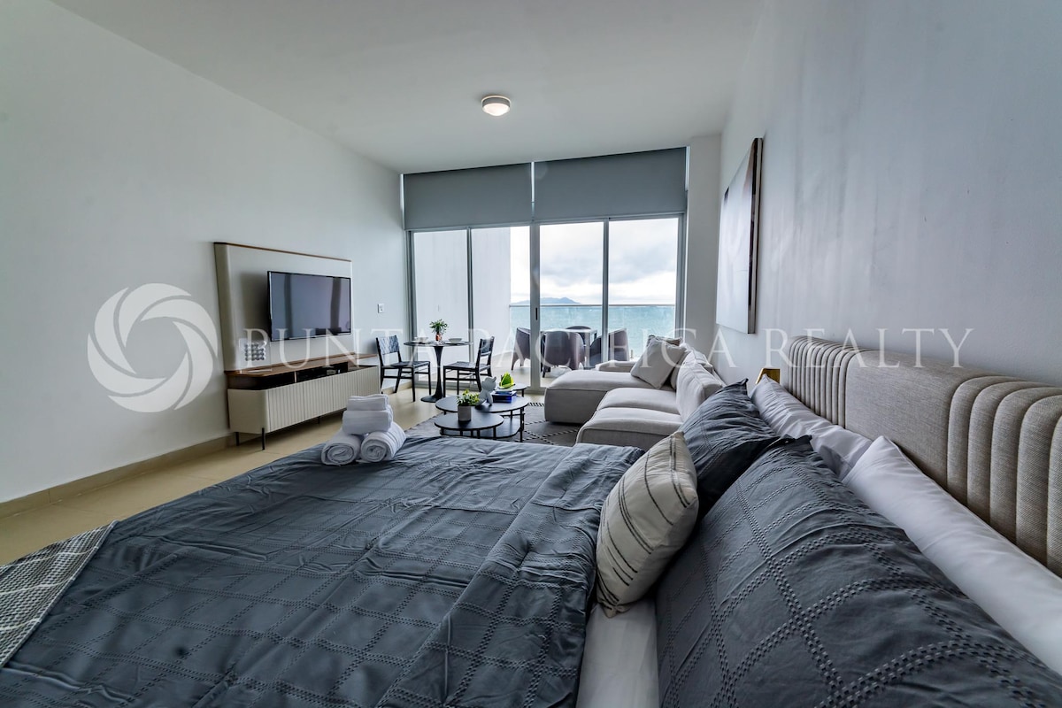 A modern studio layout is visible, featuring a spacious bed with neutral bedding. A seating area includes a sofa and table, with large windows offering views of the ocean. Soft natural light brightens the room, enhancing the clean design emphasized by muted tones.