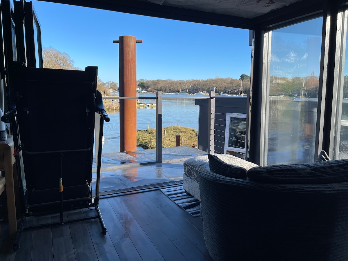 Large glass doors open to reveal views of the water and surrounding landscape. A comfortable seating area is positioned near the entrance, with sunlight illuminating the floor. A docked boat is visible in the distance, adding to the serene ambiance.