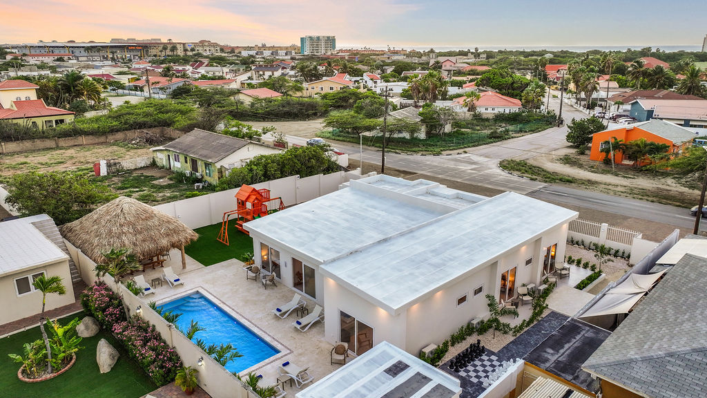 An aerial view of the villa showcases a spacious outdoor area featuring a private pool, surrounded by loungers. The landscape includes a palapa, a children's playground, and residential homes in the background, indicating a peaceful neighborhood setting.
