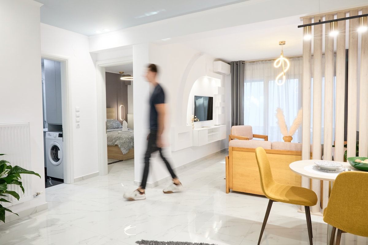 A modern living area is showcased, featuring a neutral color palette and sleek furniture. A dining table with yellow chairs is positioned near a large window, allowing natural light to brighten the space. A doorway leads to a bedroom, and a washing machine is visible in the background.