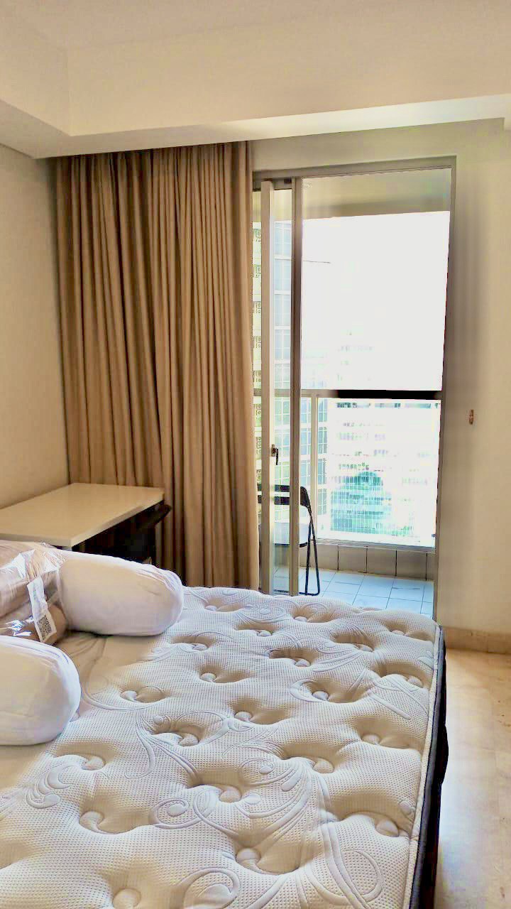 A studio bedroom features a spacious bed adorned with white pillows. Light-colored curtains frame the large sliding door leading to a balcony, allowing natural light to brighten the room. The backdrop displays a city view through the glass railing.