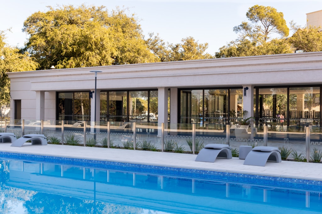 A large semi-olympic pool is framed by a spacious patio area with lounge chairs. The modern, single-story building features expansive glass windows reflecting the surrounding greenery. The peaceful atmosphere is enhanced by ample outdoor space, inviting relaxation and enjoyment of the outdoors.