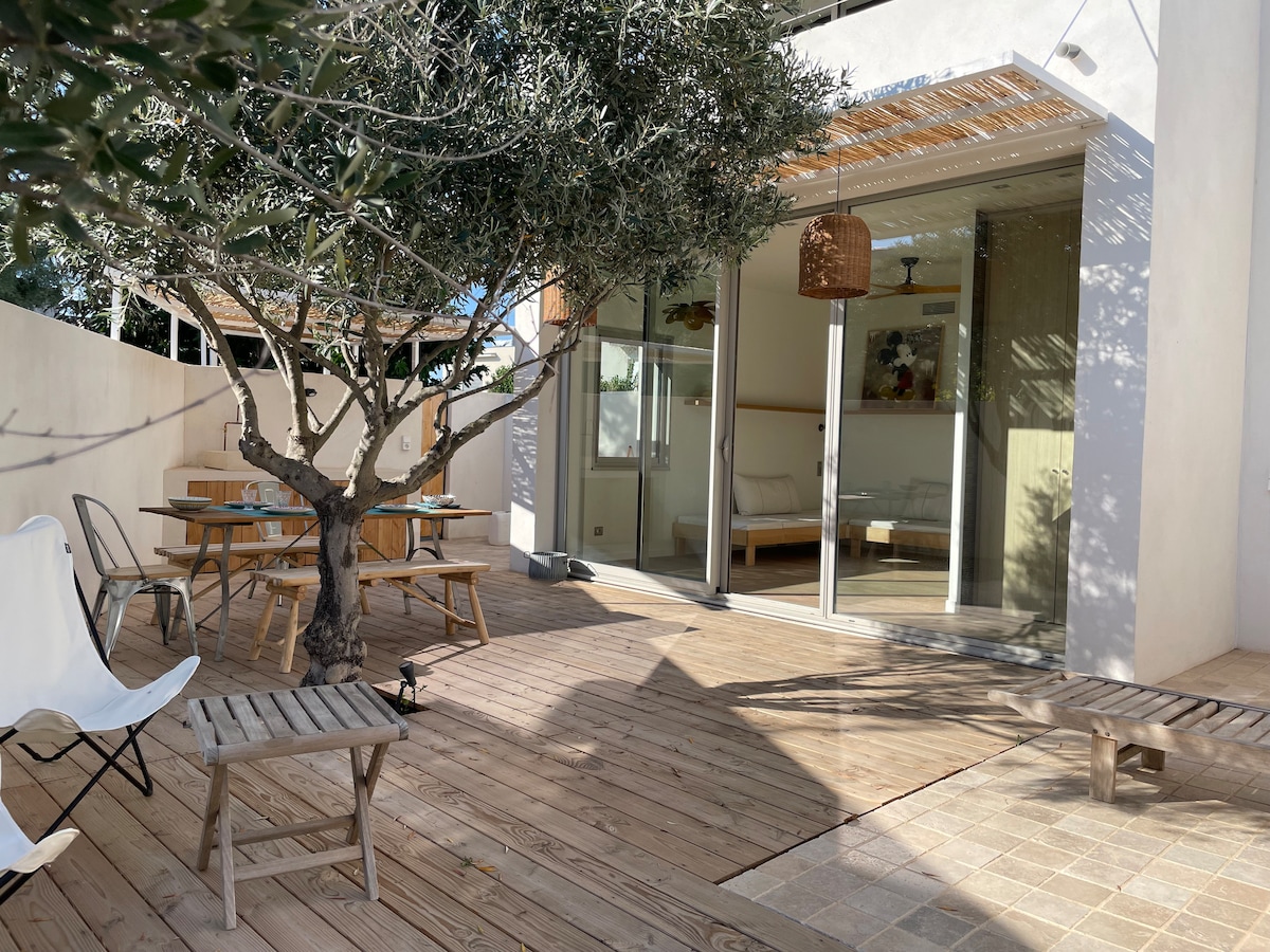 An outdoor living area is featured with wooden decking and a shaded dining space under an olive tree. A spacious glass door leads to an interior room, while a few minimalist chairs and small tables are positioned around the area.