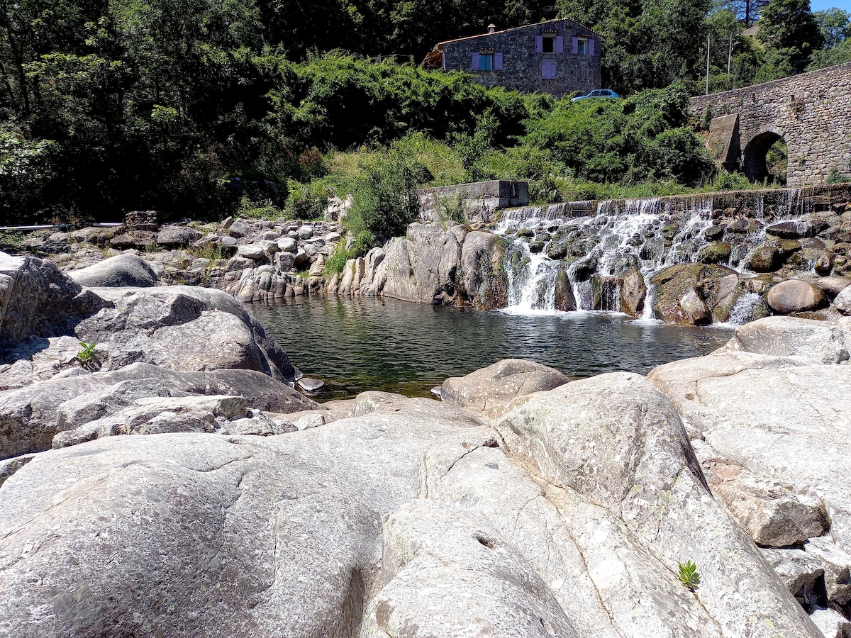 A serene river scene is depicted, featuring smooth rocks surrounding clear water. A gentle waterfall cascades over the stones, while lush greenery frames the area. In the background, a rustic building with stone details is visible, harmonizing with the natural surroundings.