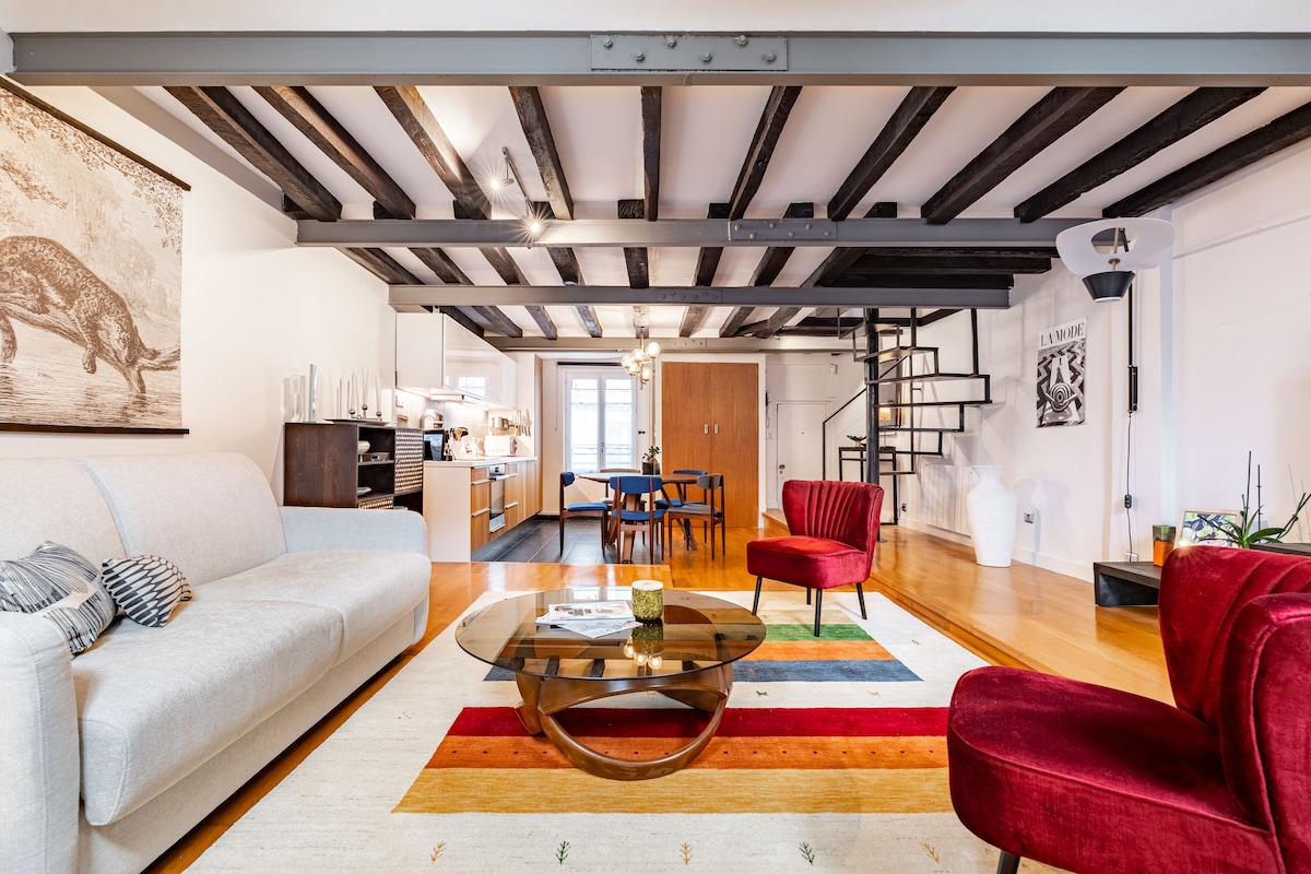 A spacious living area features a light-colored sofa and two red armchairs surrounding a round coffee table. Exposed beams are visible on the ceiling, and a colorful area rug lies underfoot, adding warmth to the modern decor. The kitchen and dining area are seen in the background.
