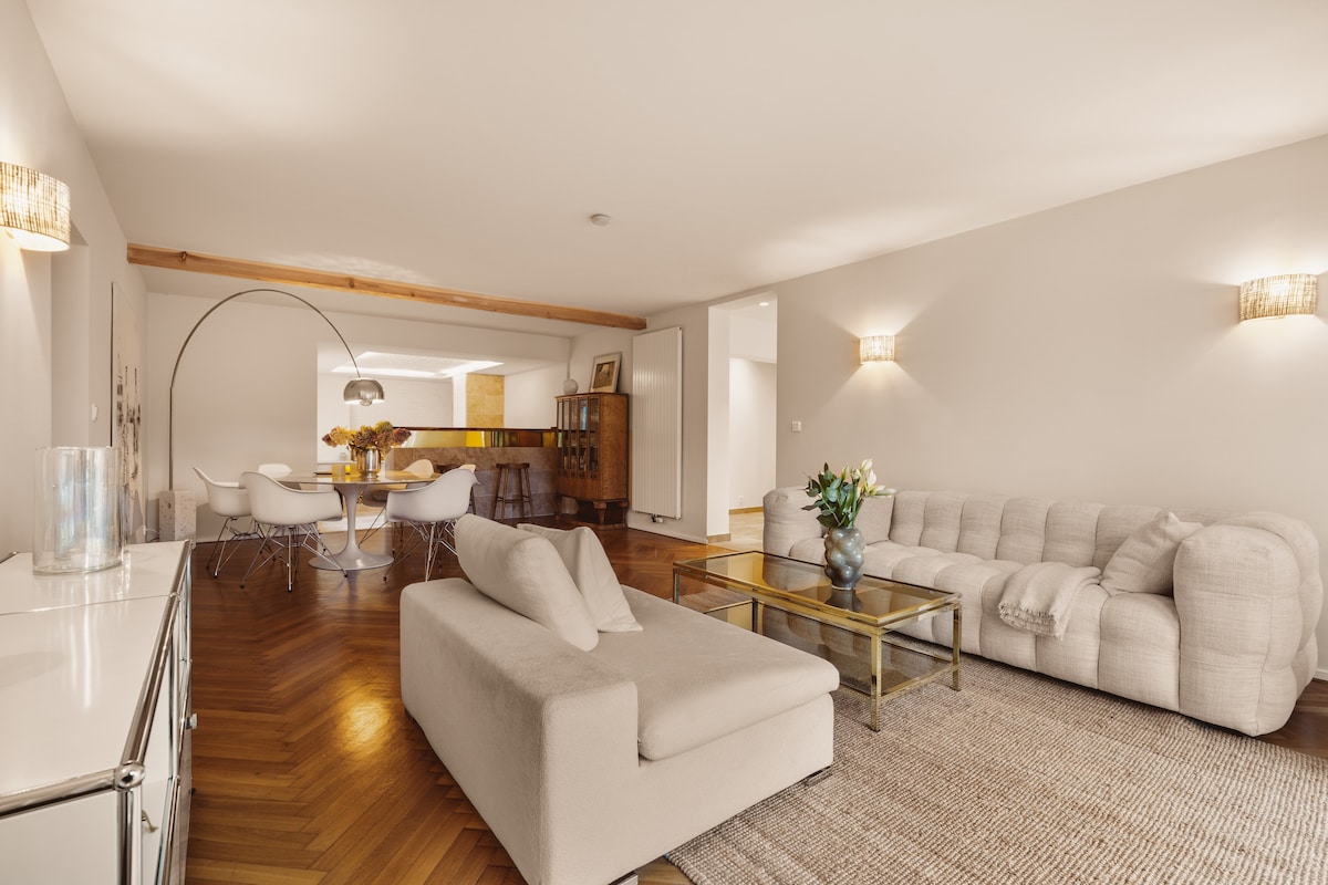 A spacious living area features a neutral-toned sofa and a glass coffee table, complemented by warm wooden flooring. Soft lighting from wall sconces illuminates the room, while a dining space with white chairs and a wooden table is visible in the background.