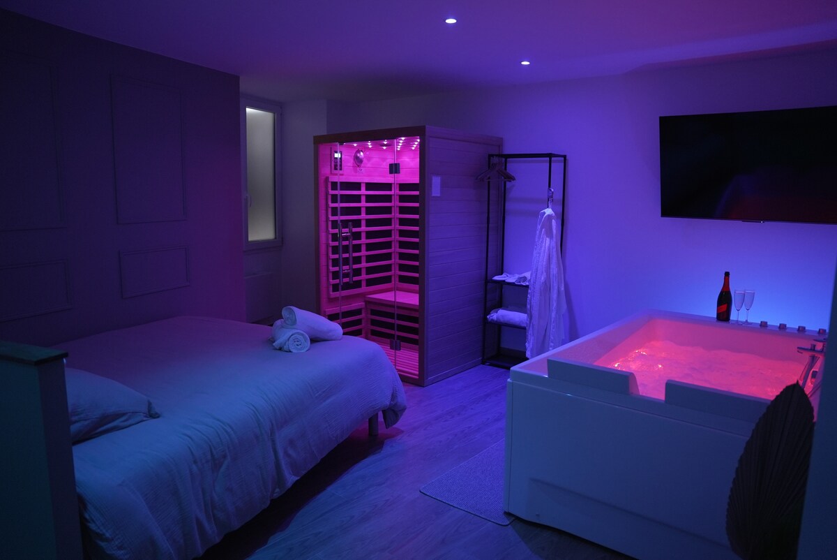 The room features a spacious bed accompanied by soft towels. A sauna with ambient lighting is positioned beside a jacuzzi, illuminated in a soothing glow. A flat-screen TV is mounted on the wall, and a white robe hangs nearby, enhancing the relaxation space.