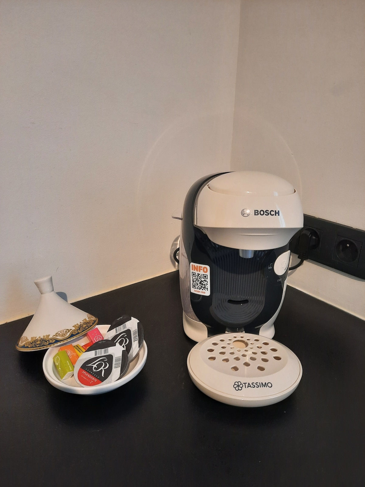 A coffee machine by Bosch is positioned on a dark countertop alongside a small, white bowl containing various coffee pods. The machine features a sleek, modern design with a QR code positioned on its front. A decorative vase is situated nearby.
