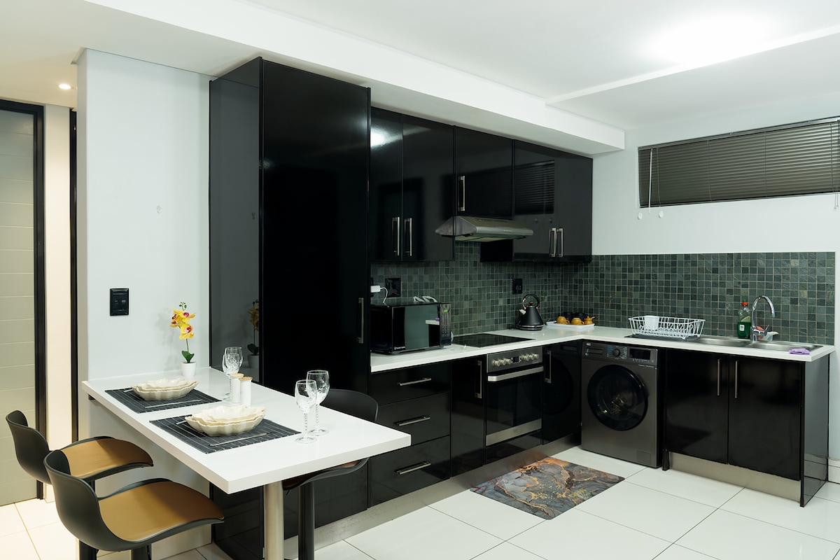 A modern kitchen is featured with sleek black cabinets and a tiled backsplash in shades of green. The space includes stainless steel appliances, a two-person dining table, and a washing machine. Natural light is provided by a window above the sink.
