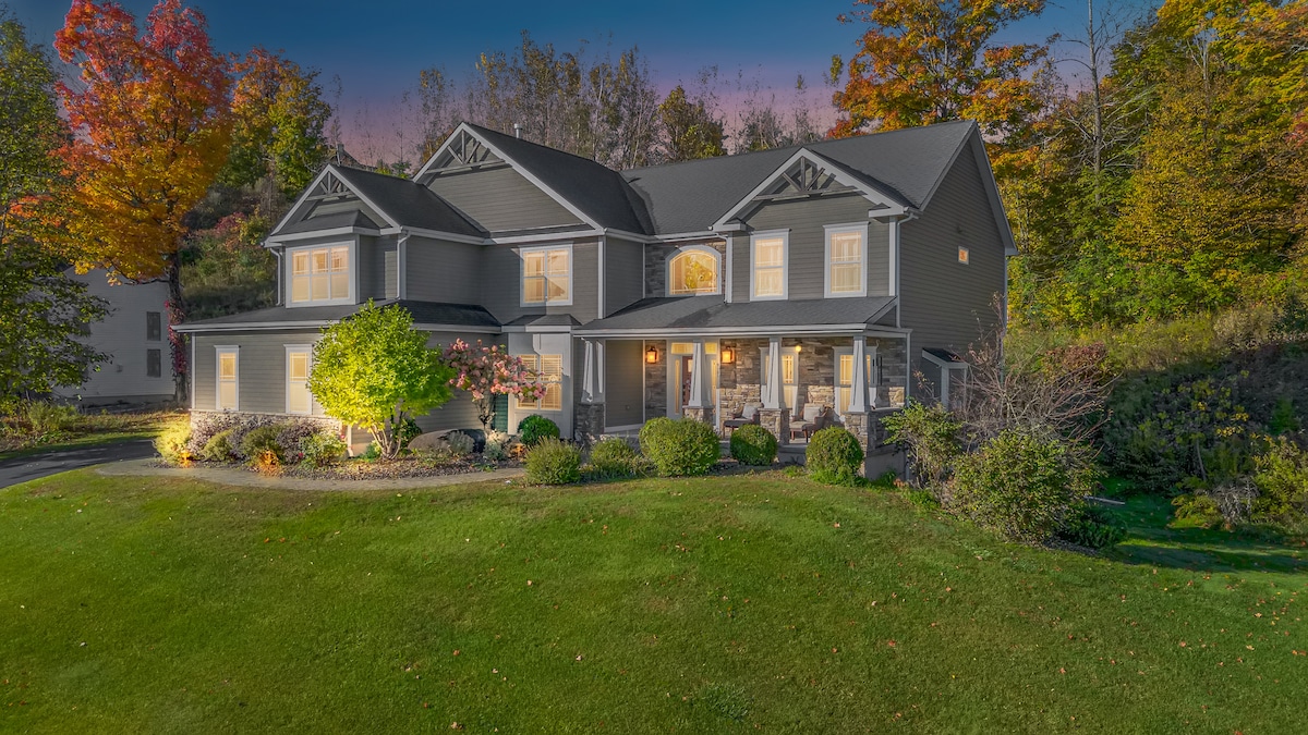 The exterior of a spacious custom-built home is presented, featuring a well-manicured lawn surrounded by vibrant autumn foliage. A welcoming front porch with seating invites relaxation, while the property is highlighted by decorative landscaping and soft evening lighting.