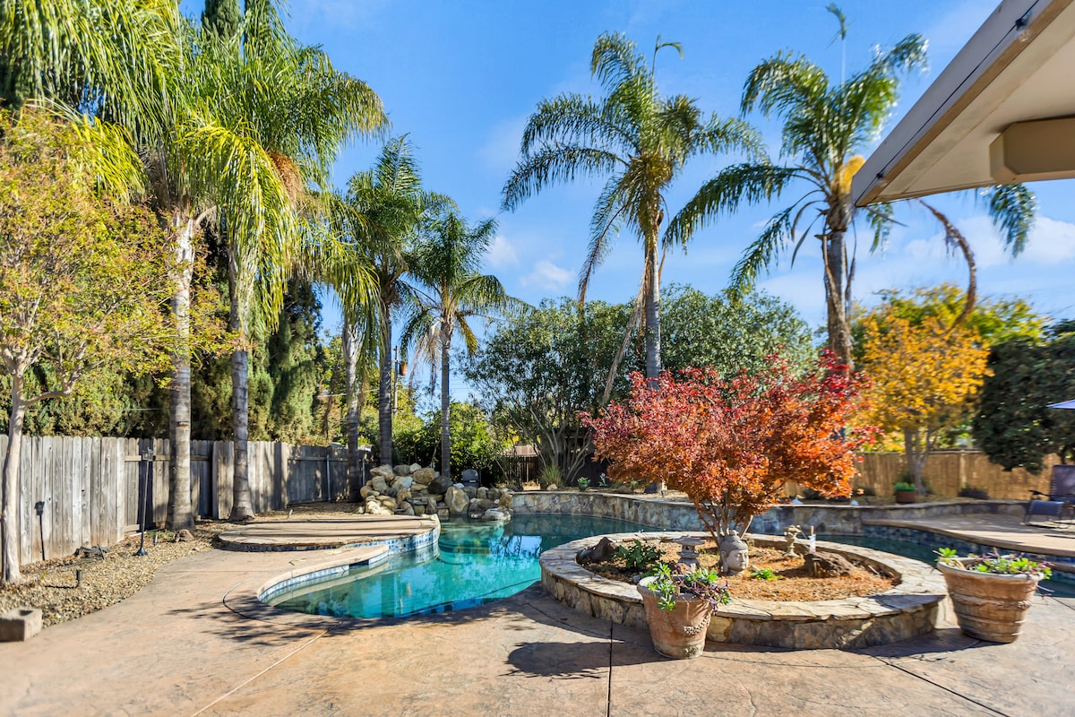 A spacious backyard features a pool surrounded by stonework and tropical palm trees. A vibrant tree with red foliage adds color to the scene. A rock waterfall flows into the pool, complemented by a stone patio, creating a relaxing outdoor environment.