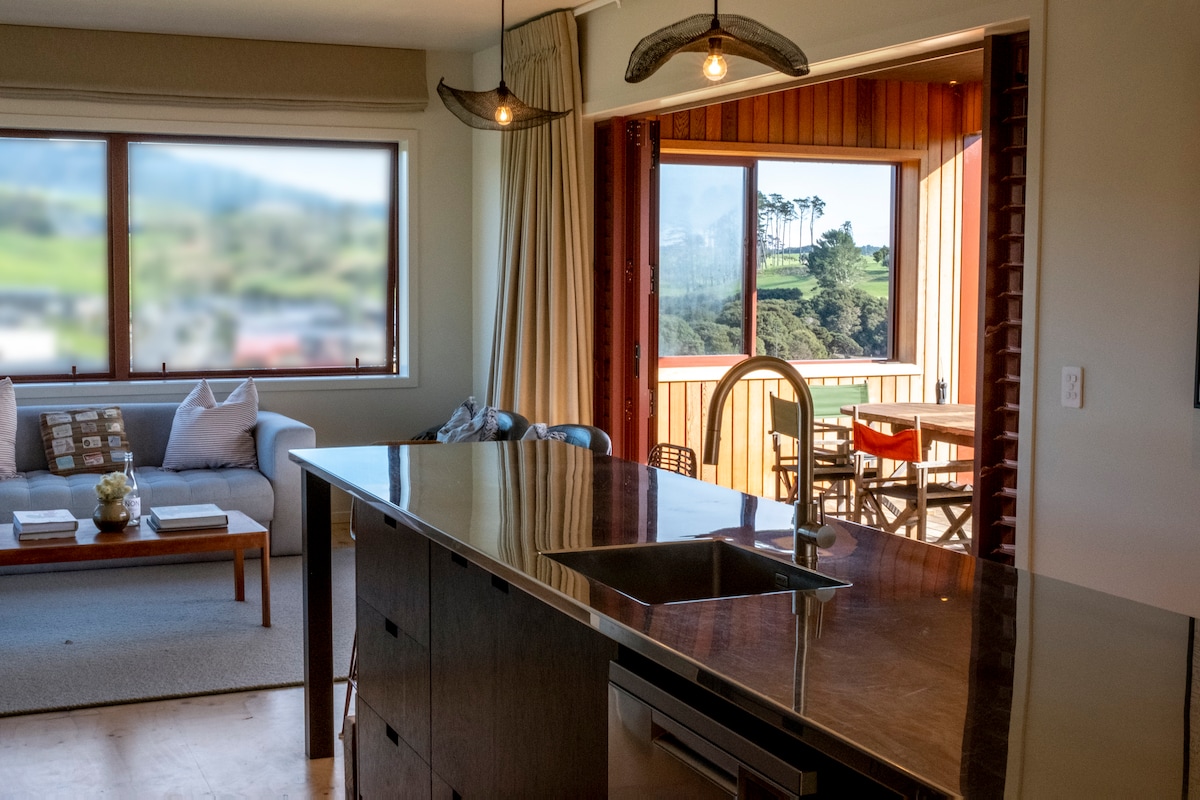 A modern kitchen area features a sleek countertop and an open view through large windows, connecting indoor and outdoor living spaces. Natural light illuminates the room, highlighting the minimalist décor and comfortable seating arrangements visible in the living area beyond.